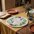 Individual circular artesanal Lilia Baez en cordón enrollado de alta densidad beige claro, con un exclusivo diseño de colibríes azules e hibiscos tropicales pintados a mano, montado en una mesa de comedor de madera clara con vajilla de terracota.