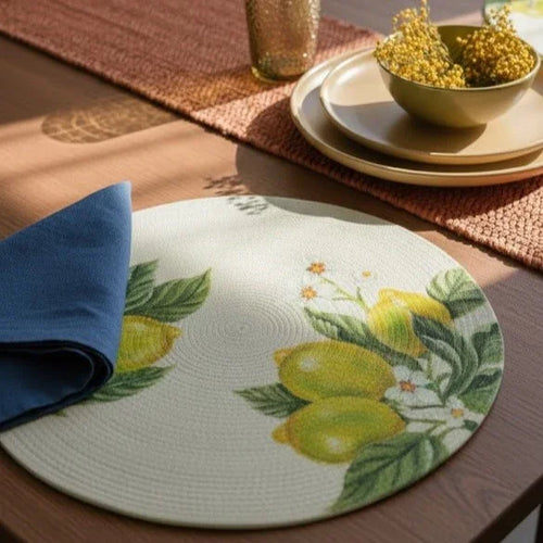 Detalle de un individual circular artesanal de diseño exclusivo Lilia Baez. El individual con limones pintados a mano está sobre una mesa de madera, con un plato beige, cubiertos y una servilleta de lino azul índigo. Fondo con un jarrón de terracota con mimosas.