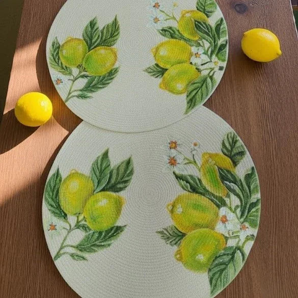 Plano cenital de dos individuales circulares grandes artesanales de diseño exclusivo Lilia Baez. El motivo es una pintura de limones y flores blancas sobre cordón beige, presentados sobre una mesa de madera con dos limones frescos enteros a los lados.