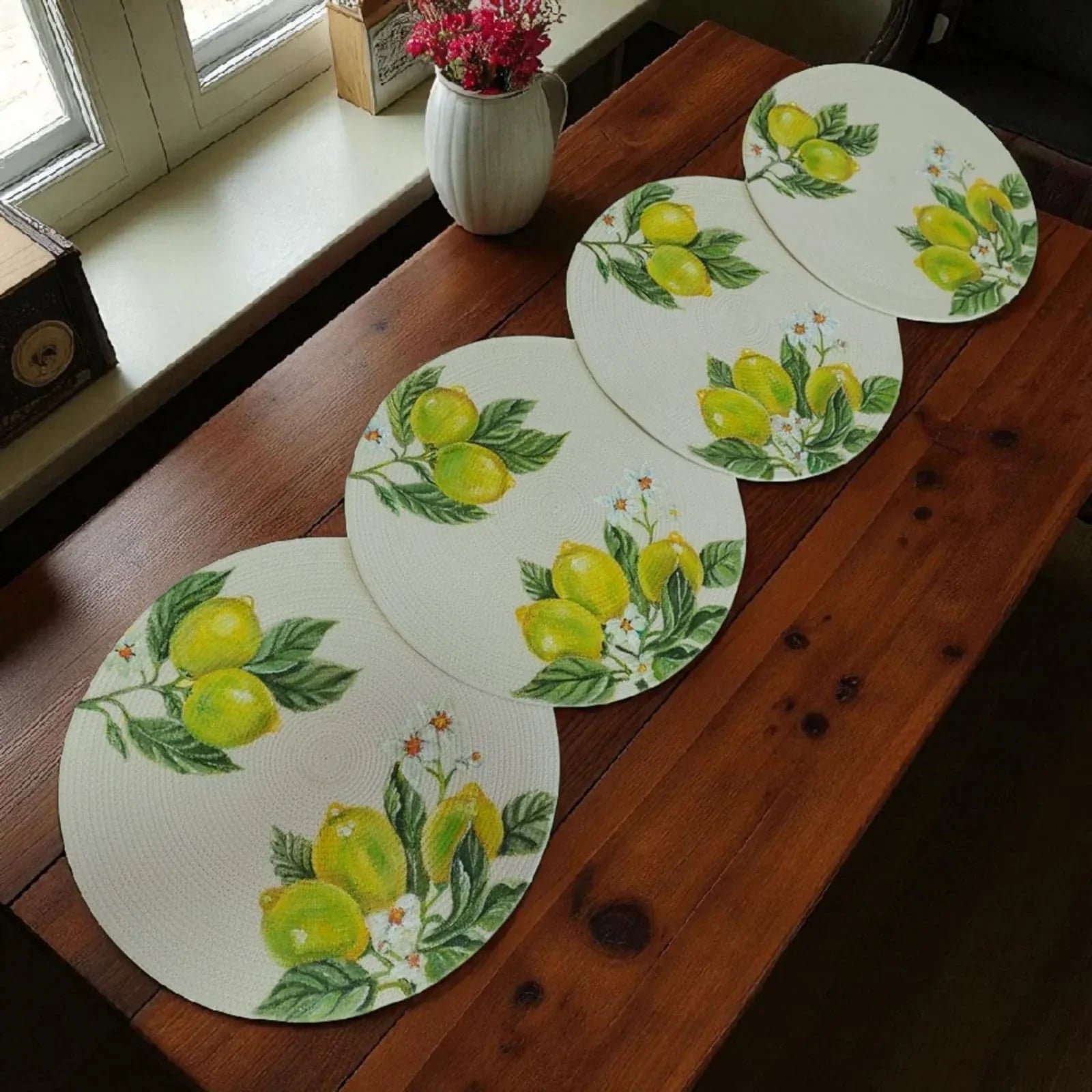 Set de cuatro individuales circulares artesanales con diseño de limones amarillos y flores blancas pintados a mano sobre cordón de alta densidad color beige claro, reposando sobre una mesa de madera texturizada.