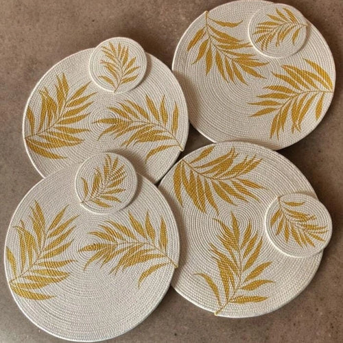 Vista cenital plana de un set de comedor artesanal Lilia Baez compuesto por cuatro individuales circulares grandes y tres portavasos circulares pequeños. Todos están elaborados en cordón enrollado de alta densidad color beige claro y presentan un diseño elegante y sofisticado de hojas de palma pintadas en oro, ideales para decoración de mesas de lujo.