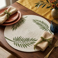Vista cenital plana de un individual circular artesanal de Lilia Baez con diseño exclusivo de tres hojas tropicales verdes pintadas a mano sobre cordón de alta densidad beige. Montado en una elegante mesa de madera con vajilla blanca, cubiertos dorados, servilleta mostaza y jarrón de terracota.